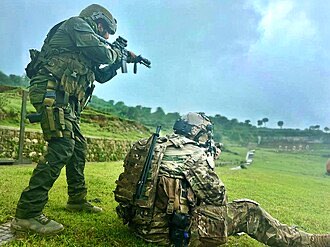 Illustrative photograph: two personnel in field clothing during a supervised training exercise on an open grass range, one standing and one kneeling.