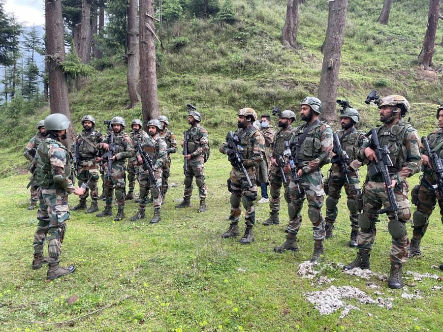 Illustrative photograph: a group of uniformed personnel gathered in a grassy clearing among tall pine trees, listening to a briefing.